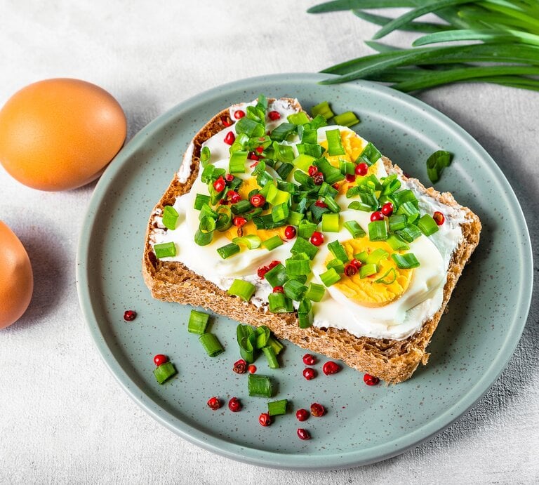 Draufsicht: Ein blauer Teller mit einem Hüttenkäse-Eier-Sandwich auf einem hellen Untergund. Daneben liegen zwei Eier mit Schale sowie grüne, schmale Blätter, wie von einer Lauchzwiebel. | © Shutterstock / irina02