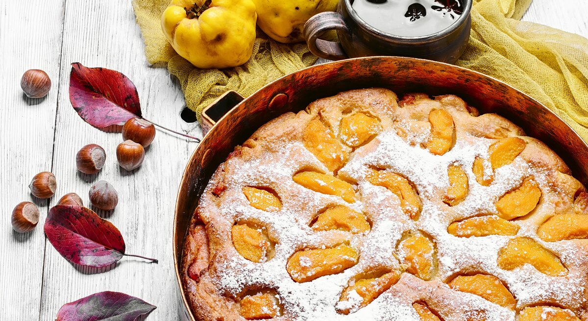 Draufsicht: ein runder Kuchen mit Quitten, der noch in der Backform ist. Er liegt auf einem hellen Untergrund aus Holz. Daneben liegen Blätter von einem Baum, Haselnüsse und Quitten. Neben dem Kuchen steht eine dunkle tasse mit einer dunklen Flüssigkeit.