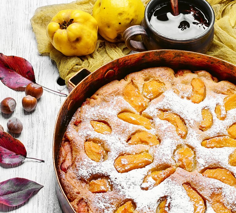 Draufsicht: ein runder Kuchen mit Quitten, der noch in der Backform ist. Er liegt auf einem hellen Untergrund aus Holz. Daneben liegen Blätter von einem Baum, Haselnüsse und Quitten. Neben dem Kuchen steht eine dunkle tasse mit einer dunklen Flüssigkeit. | © Shutterstock / Lunov Mykola