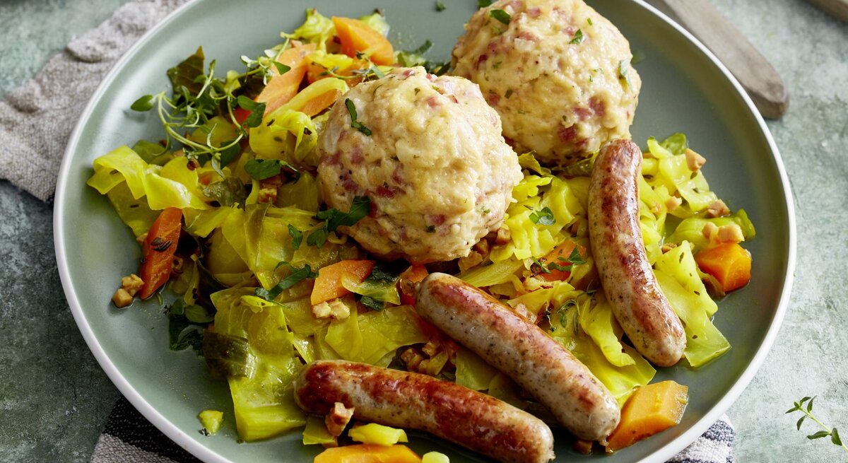 Auf einem Teller ist eine Portion Speckknödel auf geschmortem Weißkohl angerichtet. Dazu gibt es Rostbratwürstchen. 