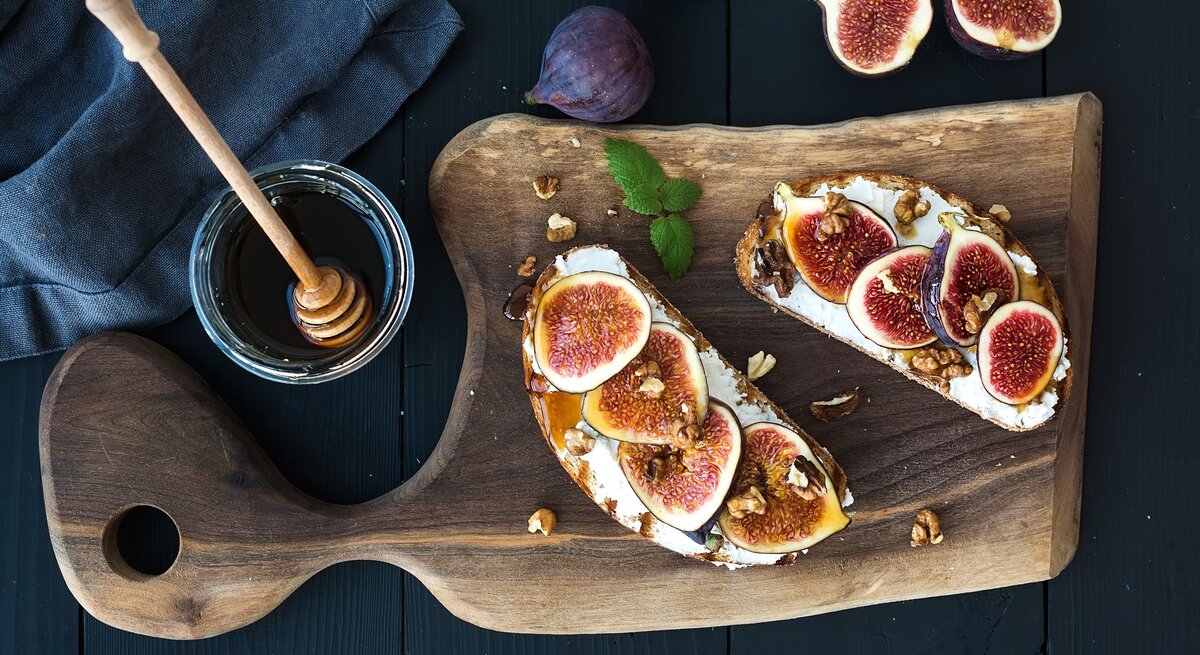 Zwei Brotscheiben mit Ricotta, Feigenund Honig auf einem dunklen Tisch. Daneben einige frische Feigen, ein Glas Honig und ein blaues Tuch.