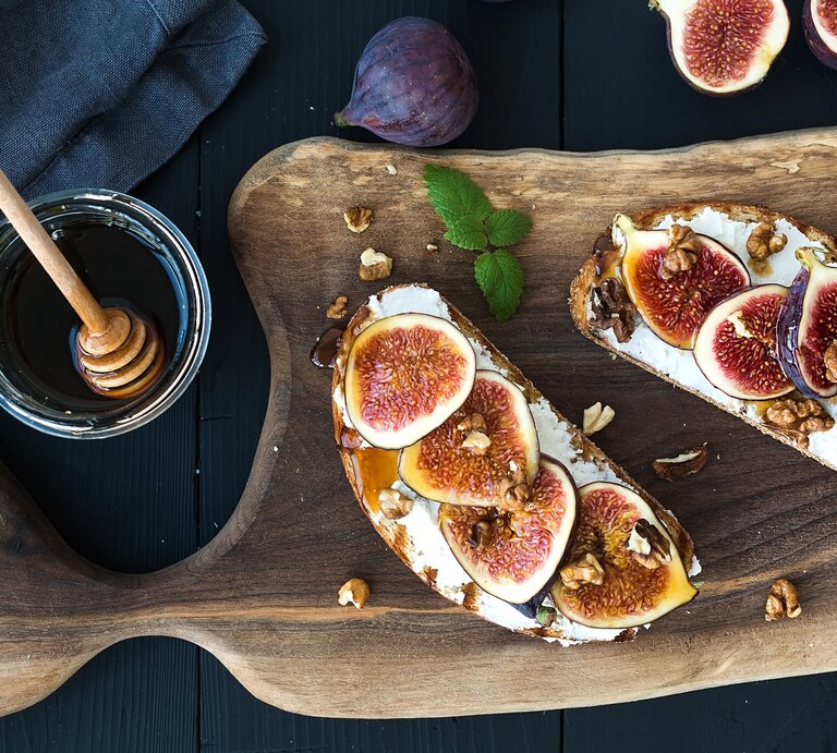 Zwei Brotscheiben mit Ricotta, Feigenund Honig auf einem dunklen Tisch. Daneben einige frische Feigen, ein Glas Honig und ein blaues Tuch. | © Shutterstock/ Foxys Forest Manufacture