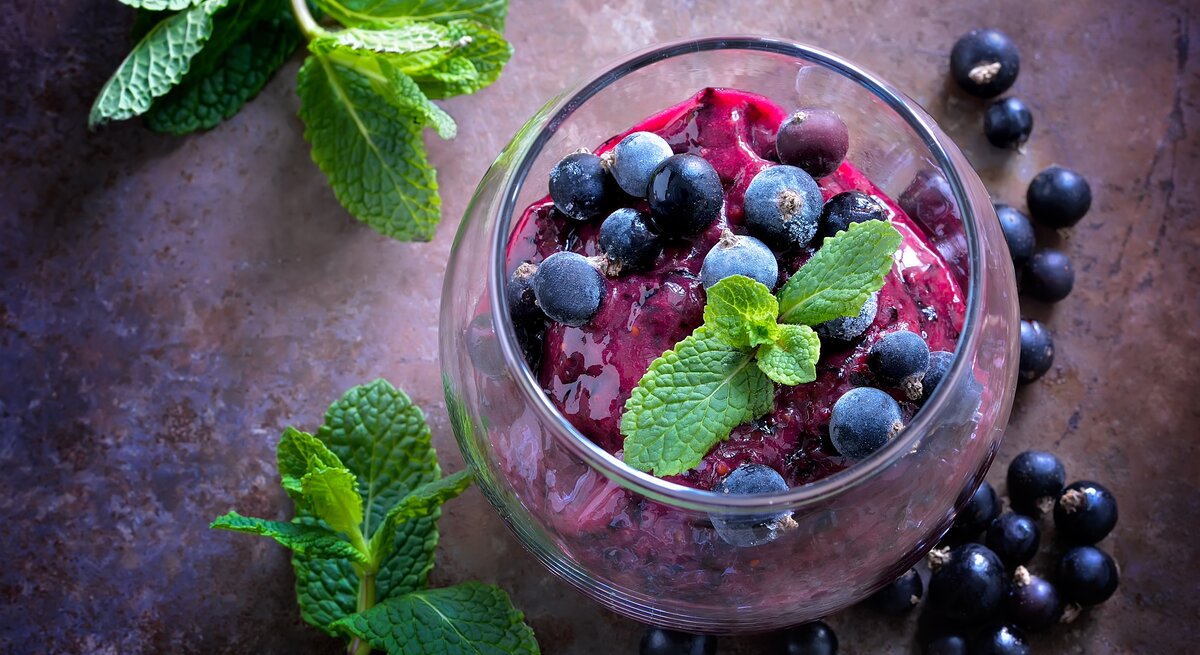 Draufsicht: Auf einem dunklem Untergrund steht ein Dessertglas mit einem Johannisbeer-Sorbet mit Prosecco. Daneben liegen schwarze Johannisbeeren sowie ein paar Blätter Minze.
