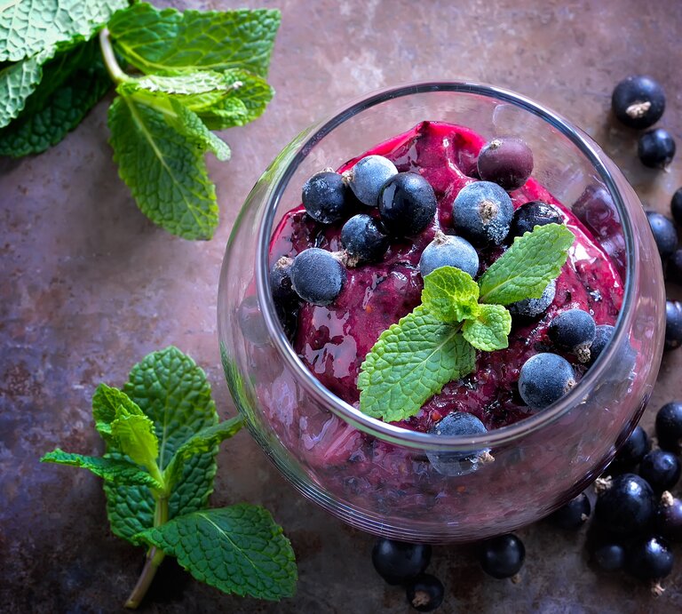 Draufsicht: Auf einem dunklem Untergrund steht ein Dessertglas mit einem Johannisbeer-Sorbet mit Prosecco. Daneben liegen schwarze Johannisbeeren sowie ein paar Blätter Minze. | © Shutterstock /  Fortyforks