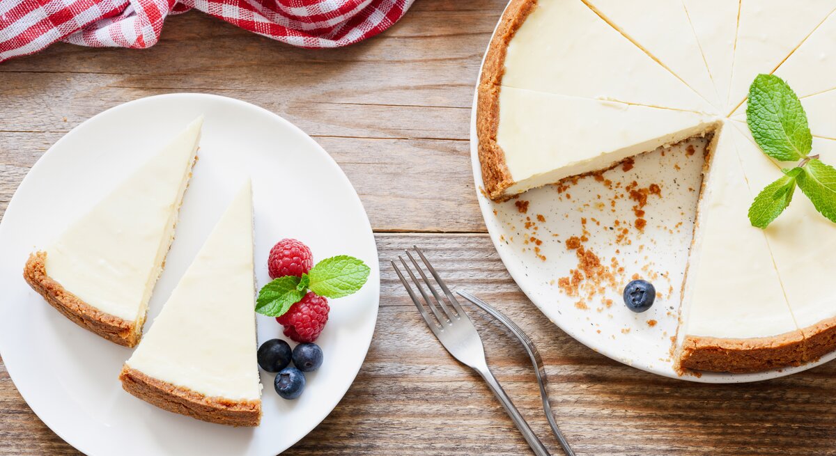 Ein Teller mit zwei Stück veganem New York Cheesecake in der Draufsicht, daneben steht der Kuchen. | © Shutterstock/ Vladislav Noseek