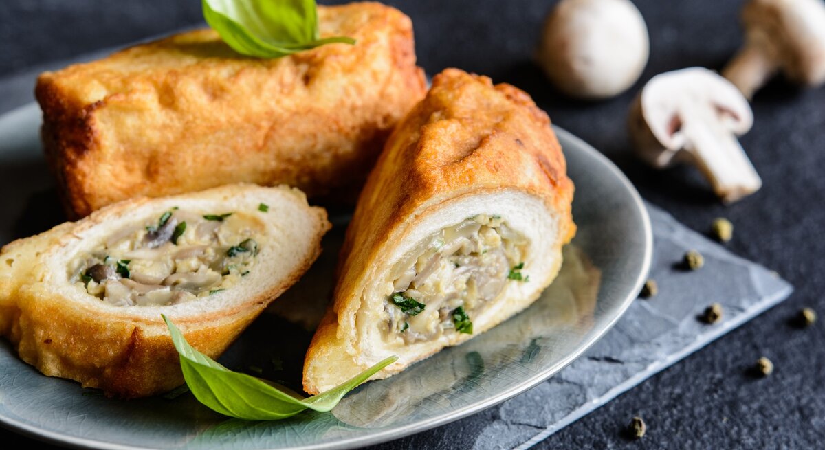 Toastrollen mit Pilzen und Käse auf grauem Teller, grauem Tuch und dunklem Tisch. Eine Rolle im Ganzen, eine aufgeschnitten dem Betrachter zugewandt. Basilikumblätter als Deko oben auf den Rollen. Im Hintergrund weiße Champignons. Frontalaufnahme.