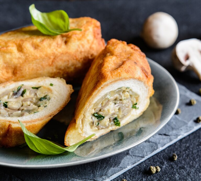 Toastrollen mit Pilzen und Käse auf grauem Teller, grauem Tuch und dunklem Tisch. Eine Rolle im Ganzen, eine aufgeschnitten dem Betrachter zugewandt. Basilikumblätter als Deko oben auf den Rollen. Im Hintergrund weiße Champignons. Frontalaufnahme. | © GettyImages/NoirChocolate