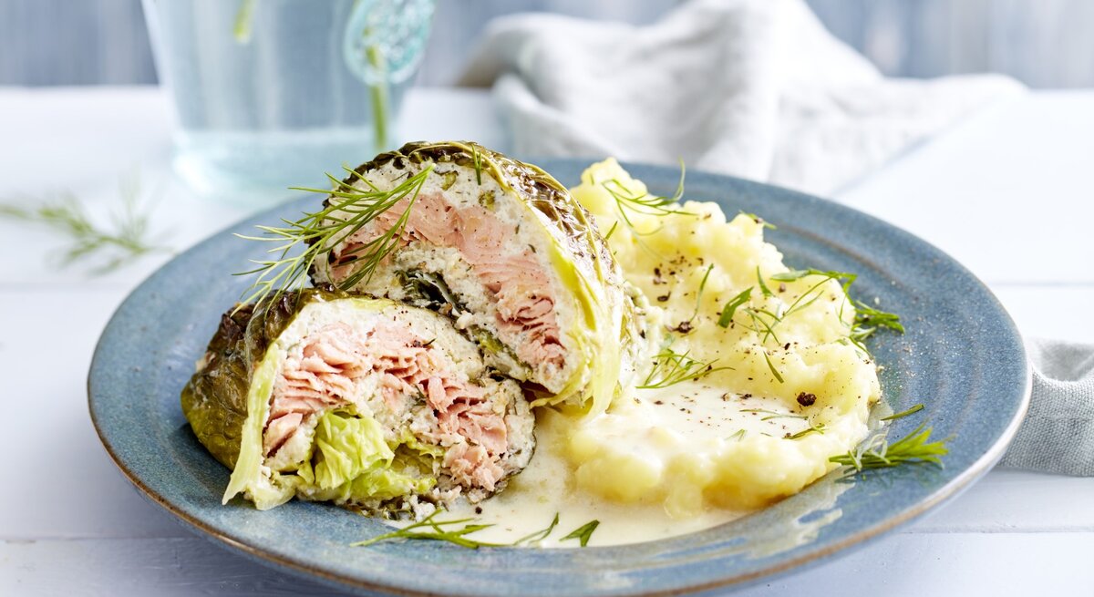 Lachs-Wirsing-Wickel, garniert mit Dill und serviert mit Kartoffelpürree auf einem Tonteller auf weiß gedecktem Tisch. 