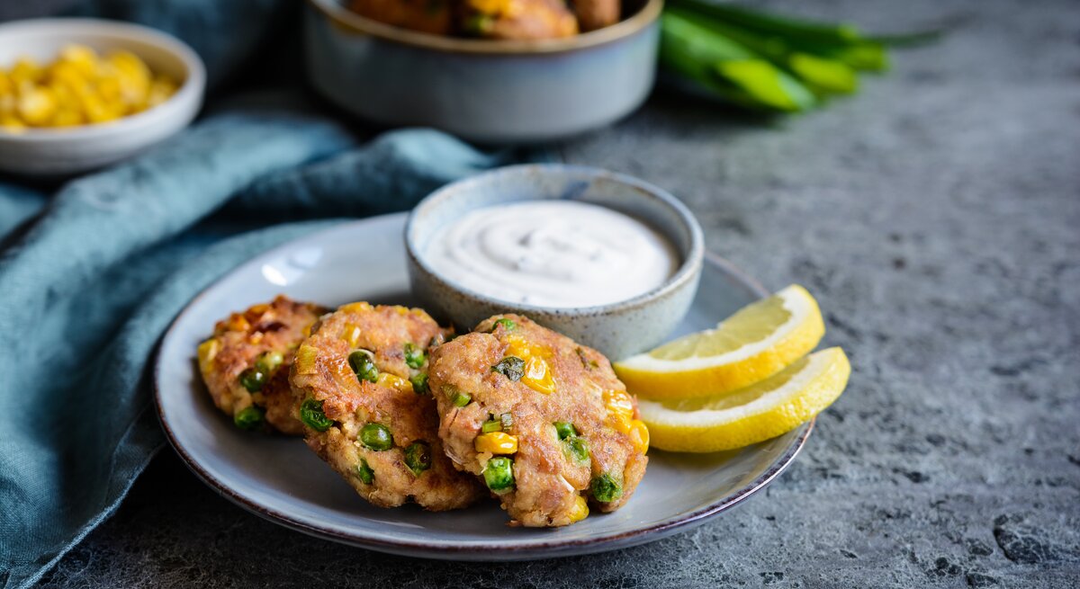 Mehrere Thunfisch-Frikadellen mit Erbsen, Mais und Frühlingszwiebeln auf einem Teller, serviert mit einem frischen Sauerrahmdip und Zitronenscheiben. 