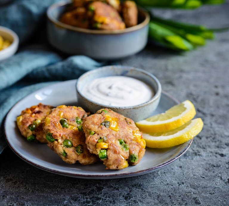 Mehrere Thunfisch-Frikadellen mit Erbsen, Mais und Frühlingszwiebeln auf einem Teller, serviert mit einem frischen Sauerrahmdip und Zitronenscheiben.  | © Getty Images / NoirChocolate