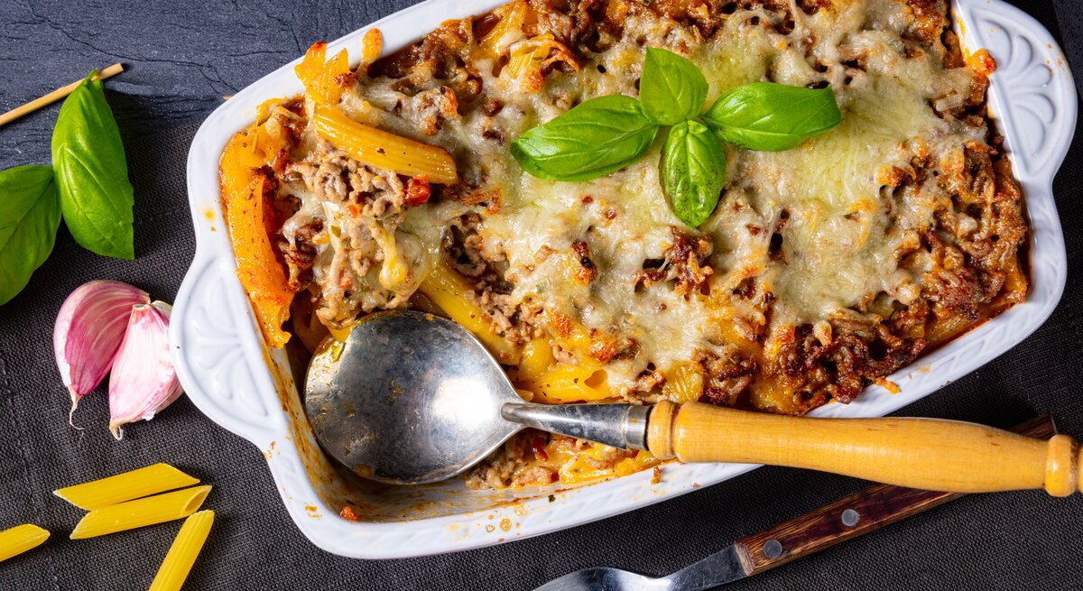 Auflauf mit Penne und Bolognese in einer Foorm, angerichtet auf dunklem Untergrund – umgeben von Basilikum, Knoblauch und Besteck.