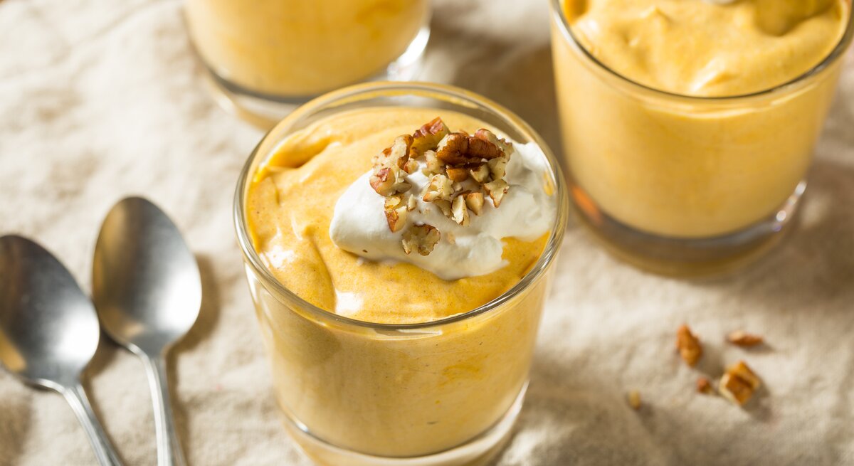 Drei Gläser gefüllt mit dem Kürbispudding der mit Sahne und Walnüssen garniert ist neben zwei Löffeln auf einer beige farbenen Tischdecke.