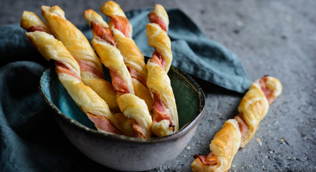 Blätterteigstangen mit Speck un einer Schüssel auf dunklem Untergrund. 