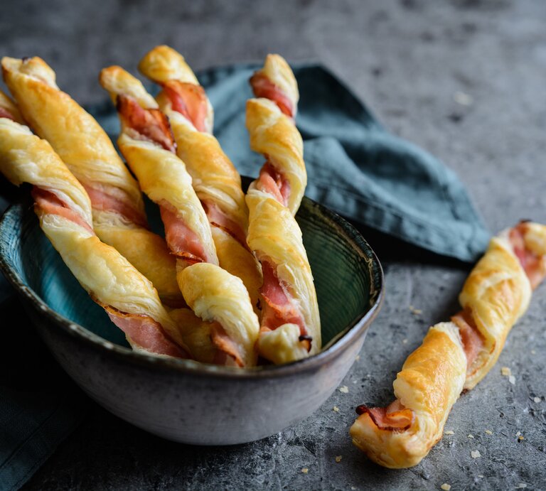 Blätterteigstangen mit Speck un einer Schüssel auf dunklem Untergrund.  | © Getty Images/NoirChocolate
