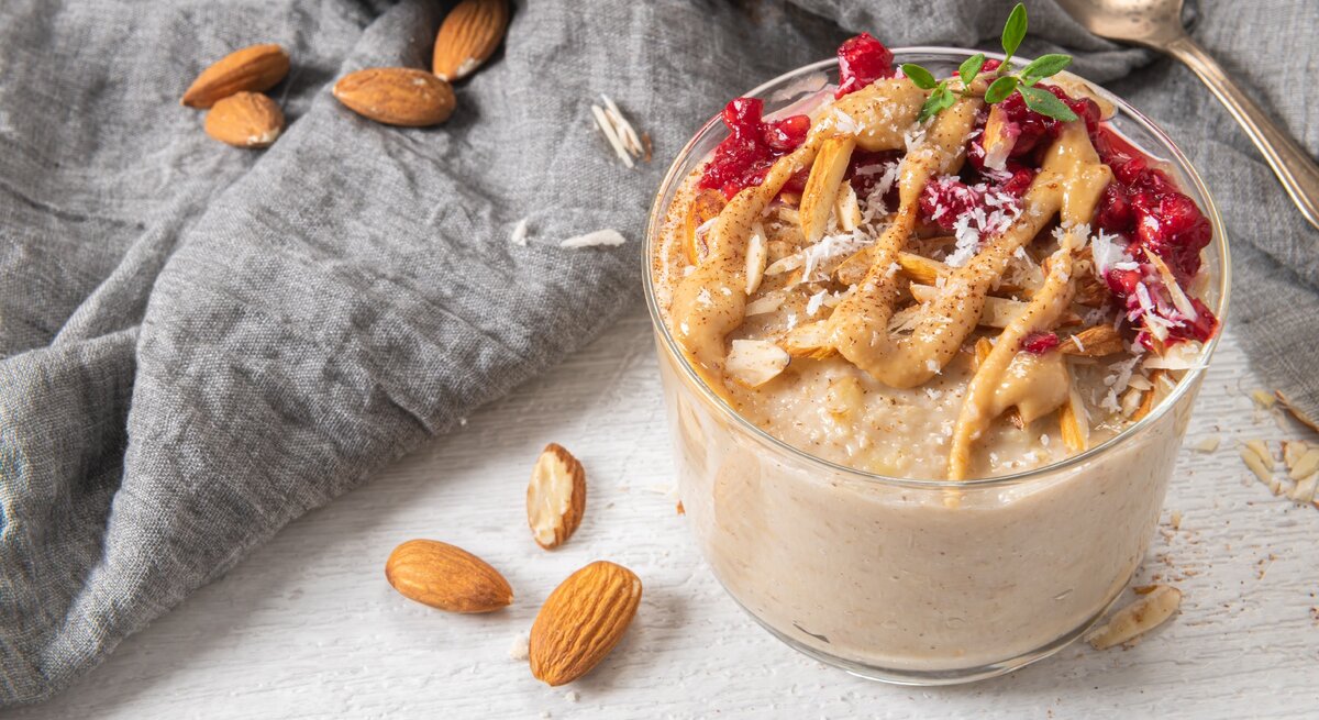 Ein Glas mit dem Mandelporridge, Himbeeren und Kokosraspeln auf einer hellen Tischplatte mit einem grauen Tuch drum herum.
