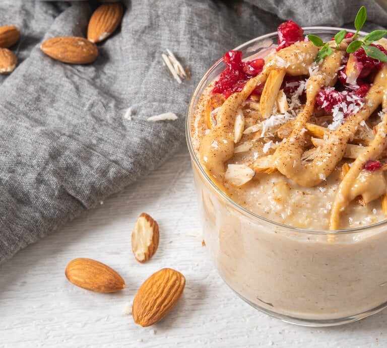 Ein Glas mit dem Mandelporridge, Himbeeren und Kokosraspeln auf einer hellen Tischplatte mit einem grauen Tuch drum herum. | © Shutterstock/ AnaMarques