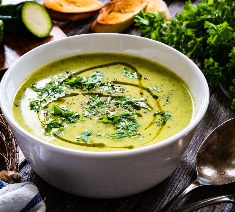 Eine Schüssel Zucchinisuppe mit Kartoffeln steht auf einem Tisch. Drumherum liegen Zucchini-Reste, Petersilie, Thymian und ein Löffel. | © Shutterstock / Chatham172