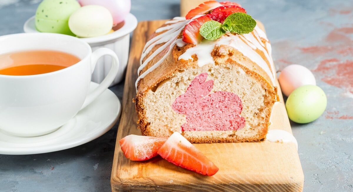 Auf einem Holzbrett liegt ein Osterhasenkuchen aus der Kastenform. Man erkennt einen rosa Hasen im Teig. Der Kuchen ist mit Zuckerguss überzogen. 