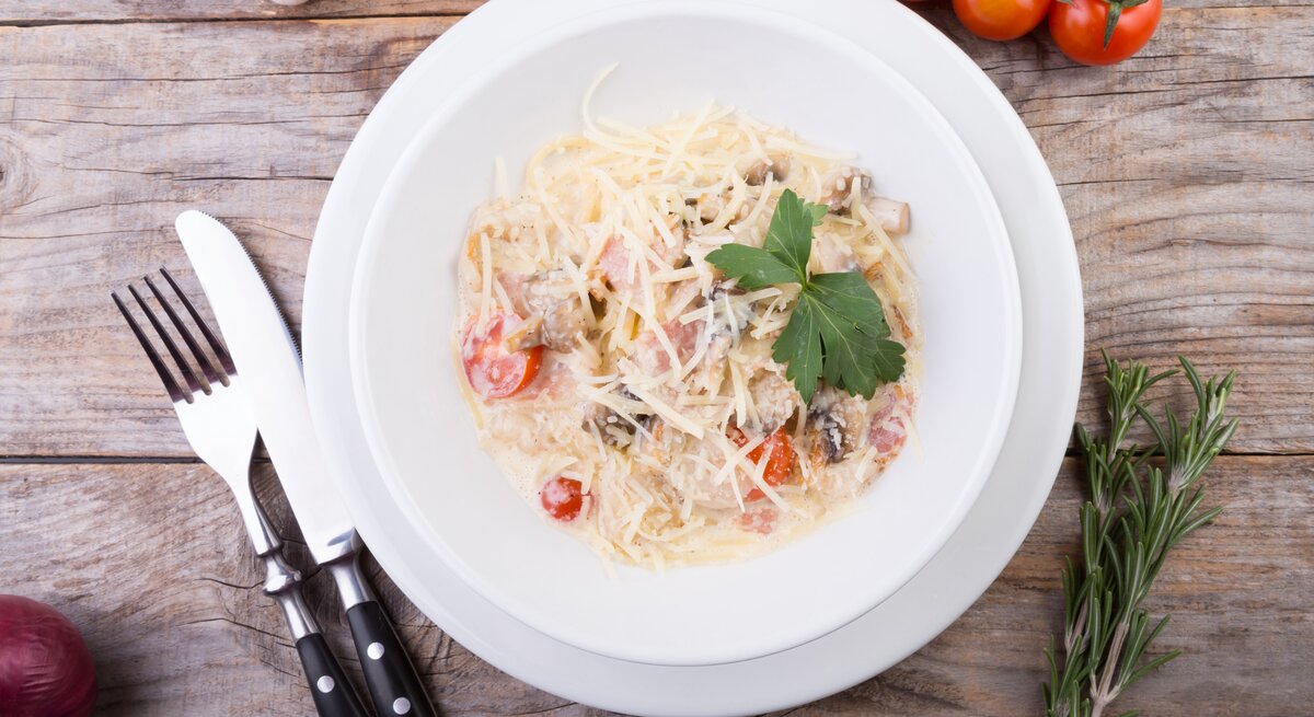 Weißer Teller belegt mit Spaghetti mit Tomaten, Pilzen und Speck in Mascarponesoße auf hölzernem Untergrund. 