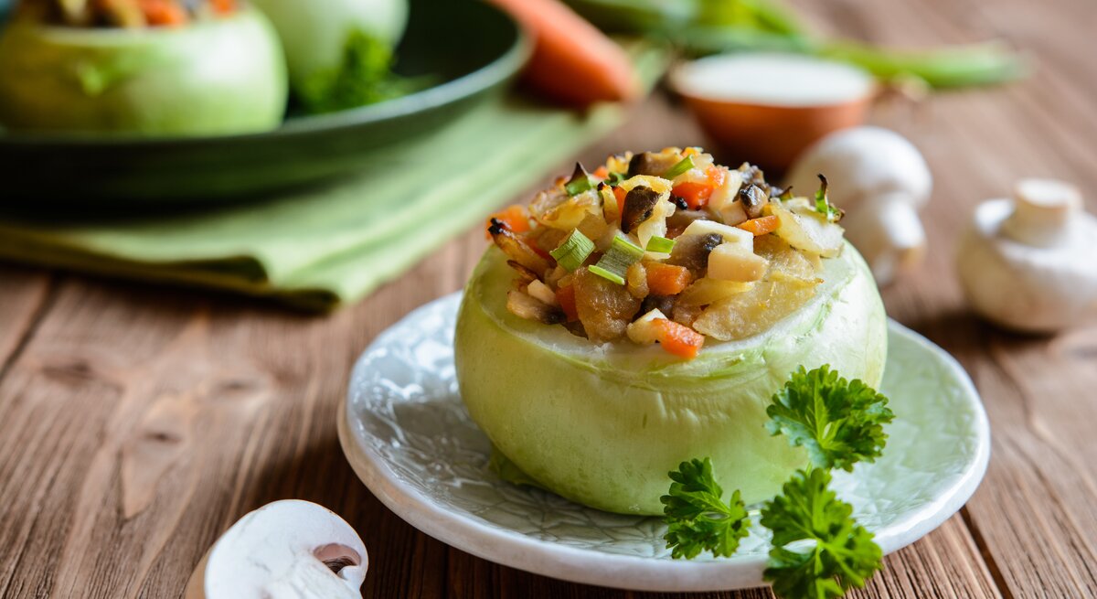 Frontalansicht: eine gefüllte Kohlrabi mit Champignons und Gemüse. Im Hintergrund sind zwei weitere Exemplare zu sehen. Drumherum liege drei frische Champignons.