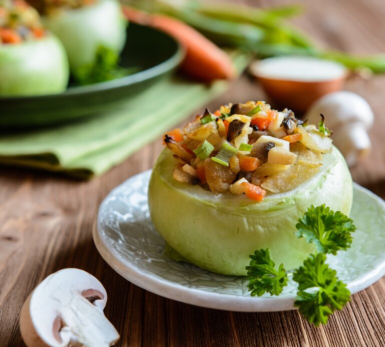 Frontalansicht: eine gefüllte Kohlrabi mit Champignons und Gemüse. Im Hintergrund sind zwei weitere Exemplare zu sehen. Drumherum liege drei frische Champignons. | © Shutterstock / NoirChocolate