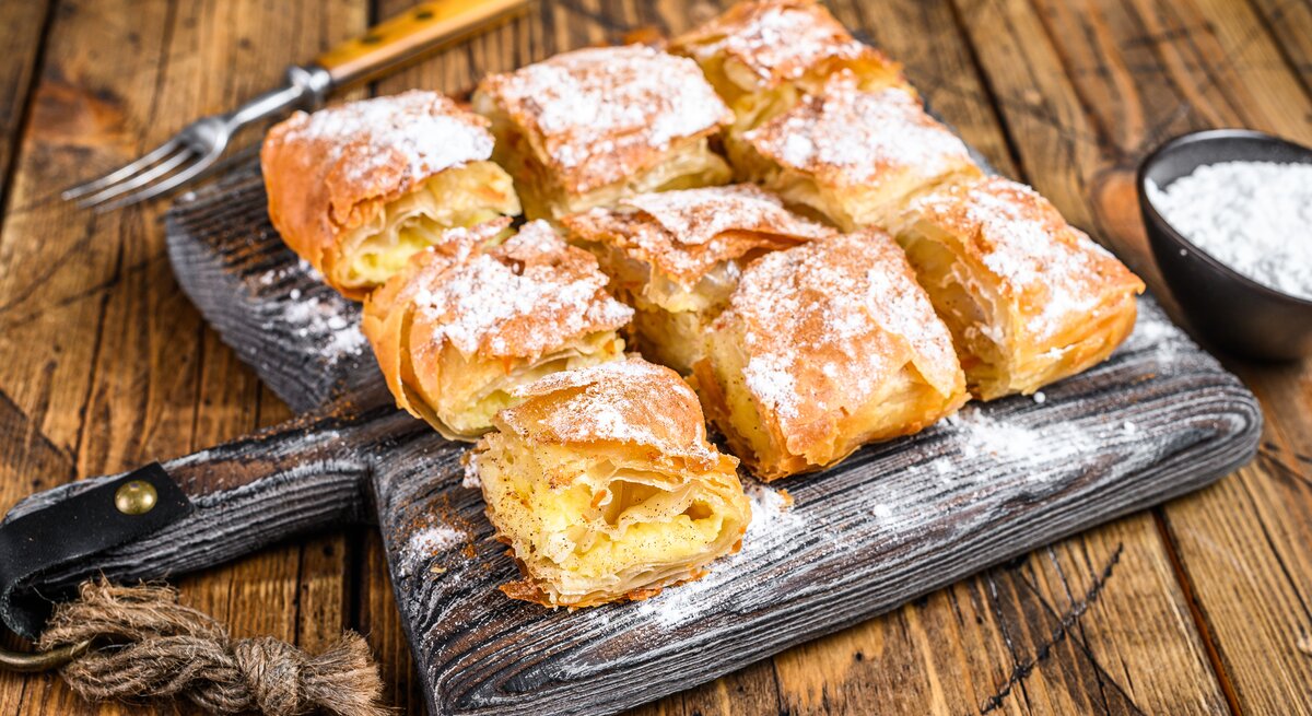 Griechischer Bougatsa aufgeschnitten auf einem schwarzen Holzbrett neben einer Gabel und einer Schüssel mit Puderzucker auf einem hellen Holztisch. 