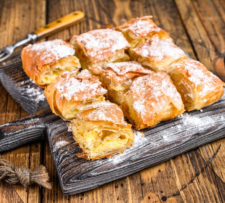 Griechischer Bougatsa aufgeschnitten auf einem schwarzen Holzbrett neben einer Gabel und einer Schüssel mit Puderzucker auf einem hellen Holztisch.  | © Getty Images / Vladimir Mironov