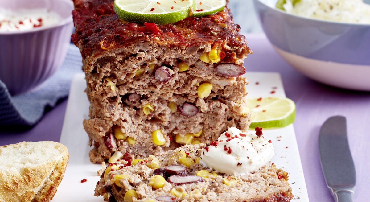 Ein tex-Mex-Braten auf einem weißen Brett. Im Hintergrund stehen zwei Schalen mit Salat und Joghurt-Dip. Der Hackbraten ist angerichtet mit Chiliflocken, Limettenscheiben und Joghurt-Dip. Neben dem Braten liegt ein Messer und zwei Scheiben Baguette.