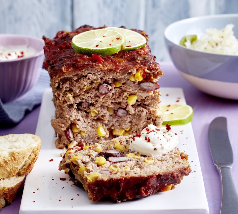 Ein tex-Mex-Braten auf einem weißen Brett. Im Hintergrund stehen zwei Schalen mit Salat und Joghurt-Dip. Der Hackbraten ist angerichtet mit Chiliflocken, Limettenscheiben und Joghurt-Dip. Neben dem Braten liegt ein Messer und zwei Scheiben Baguette. | © EAT CLUB/Isabell Triemer