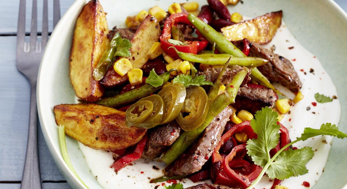 Mexikanisches Geschnetzeltes mit angerösteten Bohnen, Kartoffeln, Mais, Chili und frischem Koriander, serviert auf einem weißen Teller auf hellblauem Holztisch. 