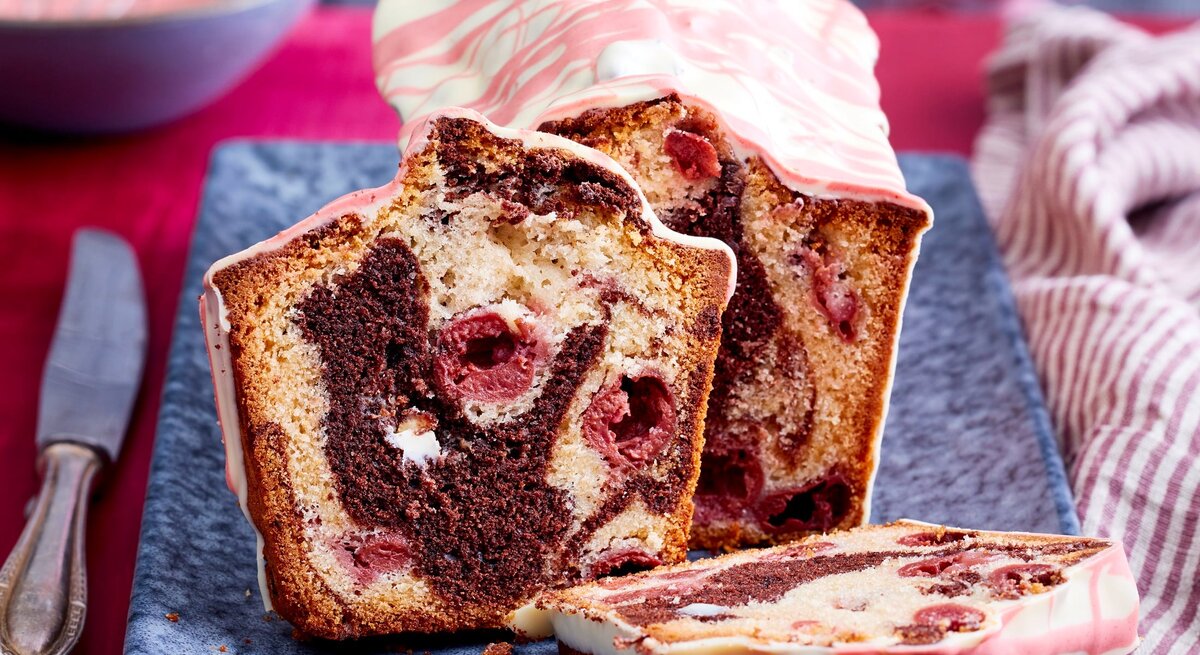 Auf einem langen Teller steht ein angeschnittener Marmor-Kirschkuchen aus der Kastenform. Er ist verziert mit einer Glasur aus weißer Schokoladen mit rosa Linien. 