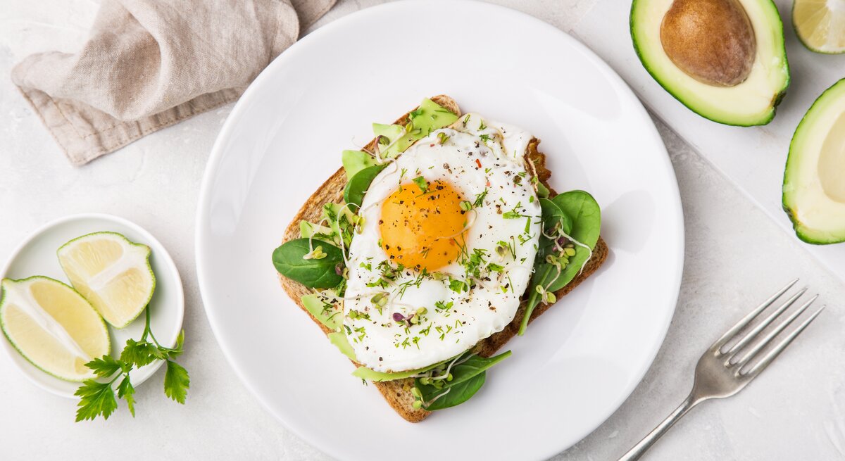 Ein Toast mit Avocado, Salat und Spiegelei liegt auf einem weißen Teller auf einem hellen Tisch. Rechts neben dem Teller liegt eine halbierte Avocado und eine Gabel. Links neben dem Teller steht eine kleine Schale mit Limettenstücken. Am oberen Bildrand liegt ein beiges Tuch.