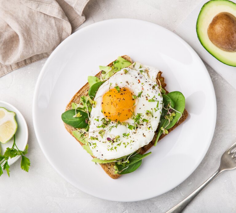 Ein Toast mit Avocado, Salat und Spiegelei liegt auf einem weißen Teller auf einem hellen Tisch. Rechts neben dem Teller liegt eine halbierte Avocado und eine Gabel. Links neben dem Teller steht eine kleine Schale mit Limettenstücken. Am oberen Bildrand liegt ein beiges Tuch. | © GettyImages / Anna_Shepulova