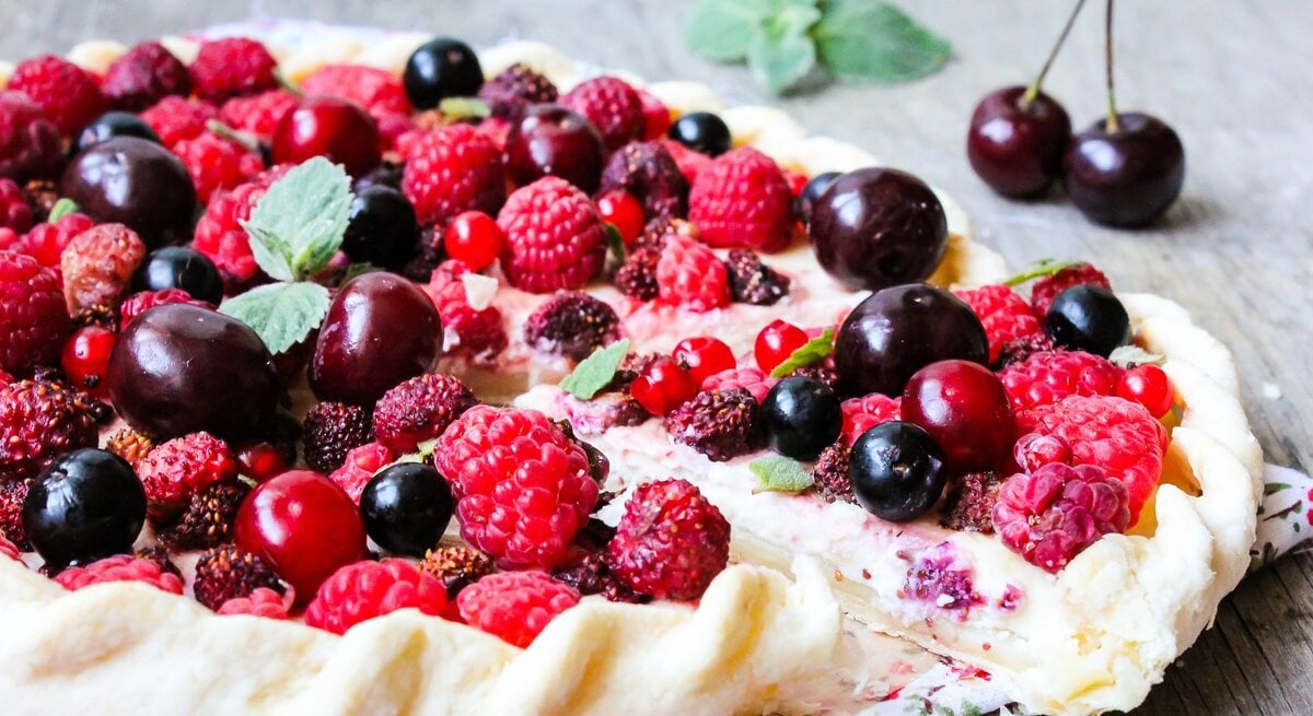 Eine Tarte mit Früchten auf einem Holzuntergrund. Im Hintergund ein Minzzweig und eine Kirsche.