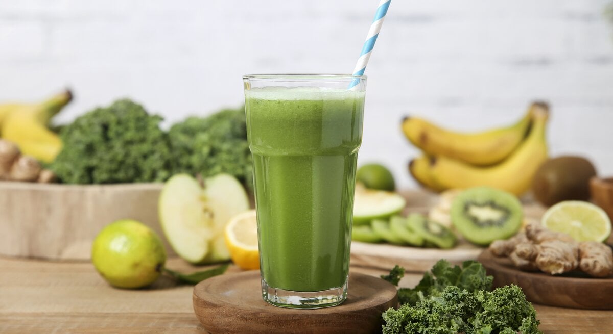 Ein Grünkohl-Smoothie in einem hohen Glas mit einem gestreiften Stohhalm. Im Hintergrund liegen Bananen, Äpfel, Grünkohl, Kiwis und Ingwer.