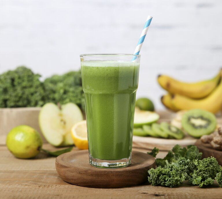 Ein Grünkohl-Smoothie in einem hohen Glas mit einem gestreiften Stohhalm. Im Hintergrund liegen Bananen, Äpfel, Grünkohl, Kiwis und Ingwer. | © Getty Images / Westend61