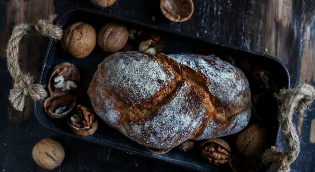 Draufsicht: Auf einem dunklen Untergrund aus Holz liegt ein Walnussbrot in einem dunklen Korb. Drumherum liegen intakte sowie geöffnete Walnüsse.
