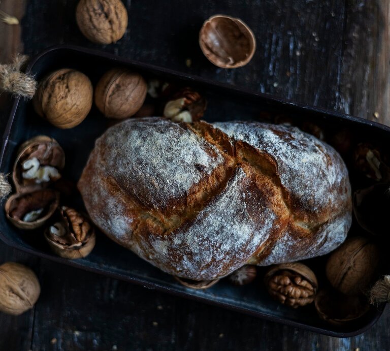 Draufsicht: Auf einem dunklen Untergrund aus Holz liegt ein Walnussbrot in einem dunklen Korb. Drumherum liegen intakte sowie geöffnete Walnüsse. | © Shutterstock / yipengge