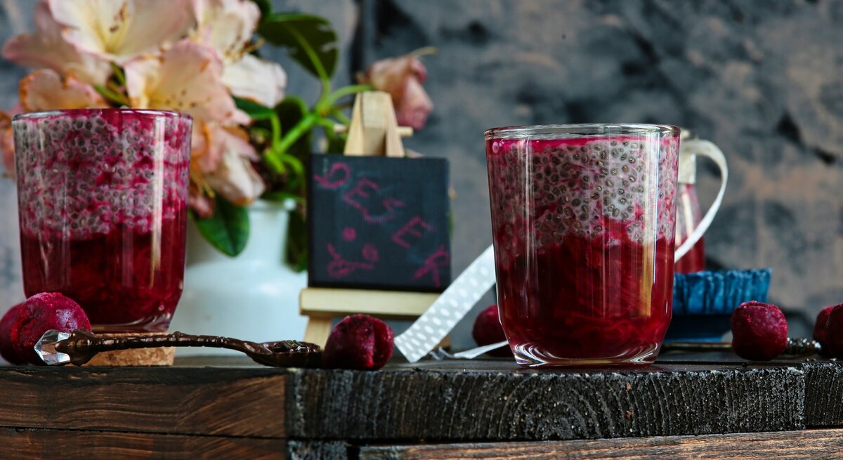 Zwei Gläser mit dem Chia-Pudding und rotem Fruchtragout auf einem Holztisch mit allerhand Deko, wie einer kleinen Tafel und ein paar Blumen.