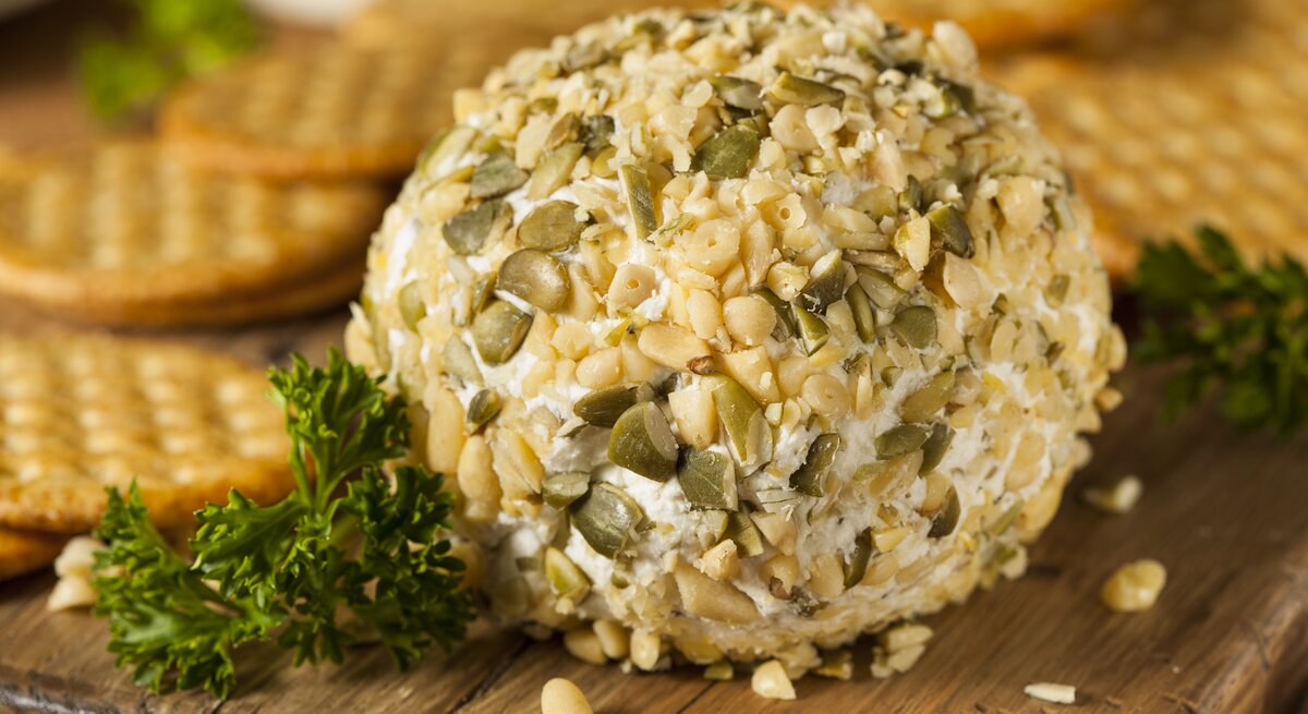 Ein Holzbrett auf dem eine Frischkäse-Kugel mit Pinienkernen und Petersilie liegt, im Hintergrund sind Cracker zu erkennen. 
