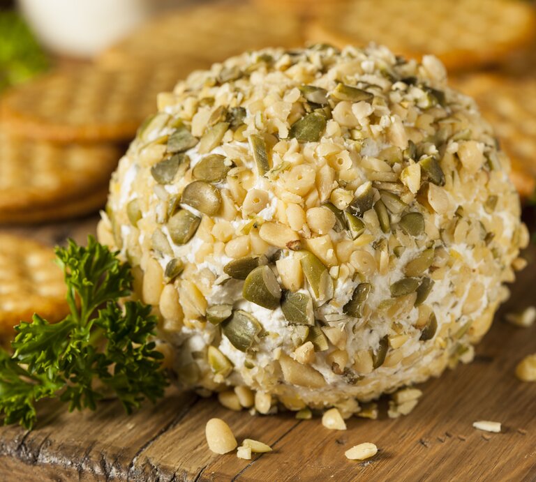 Ein Holzbrett auf dem eine Frischkäse-Kugel mit Pinienkernen und Petersilie liegt, im Hintergrund sind Cracker zu erkennen.  | © Getty Images/ bhofack2
