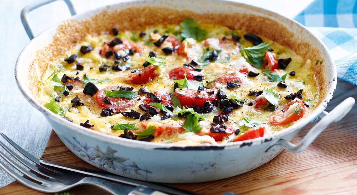 Eine Tomaten-Mozzarella-Tortilla mit schwarzen Oliven in einer Pfanne auf einem Brett, daneben Besteck.