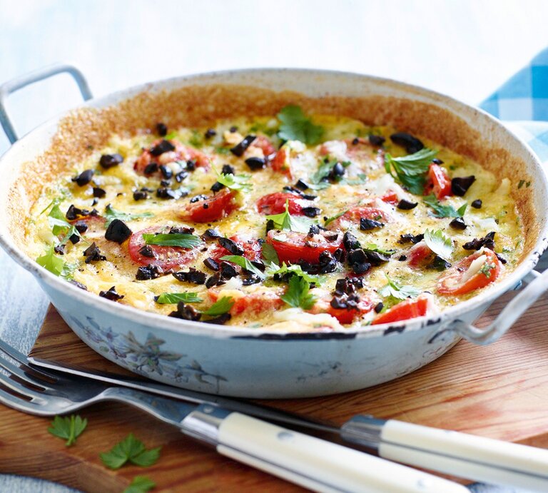 Eine Tomaten-Mozzarella-Tortilla mit schwarzen Oliven in einer Pfanne auf einem Brett, daneben Besteck. | © Becker Joest Volk Verlag/ Oliver Brachat