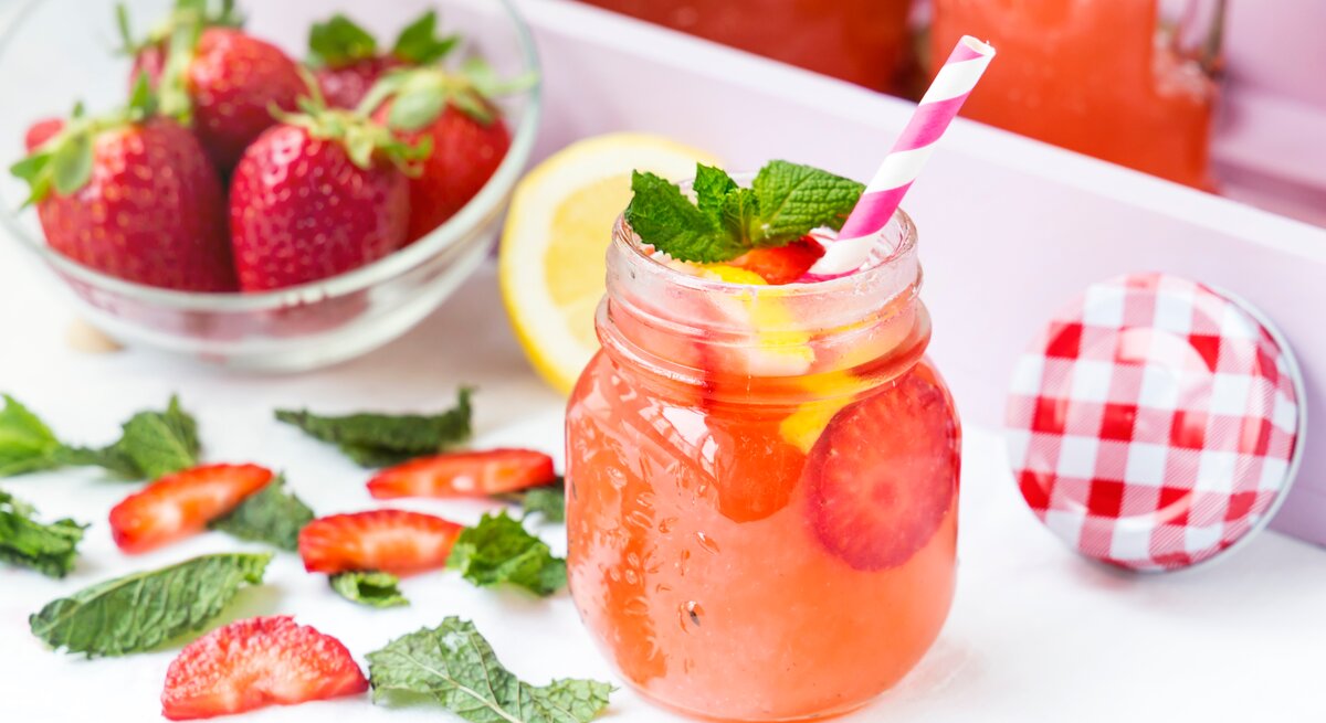 Ein alkoholfreier Erdbeer-Drink im Retro-Glas steht auf einem hellem Tablett. Drum herum liegen Erdbeere-Scheiben, eine weiße Schale mit Erdbeeren, Minzblätter sowie ein weiß-rot-karierter Deckel.