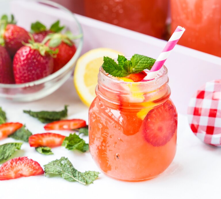 Ein alkoholfreier Erdbeer-Drink im Retro-Glas steht auf einem hellem Tablett. Drum herum liegen Erdbeere-Scheiben, eine weiße Schale mit Erdbeeren, Minzblätter sowie ein weiß-rot-karierter Deckel. | © Shutterstock / aidart