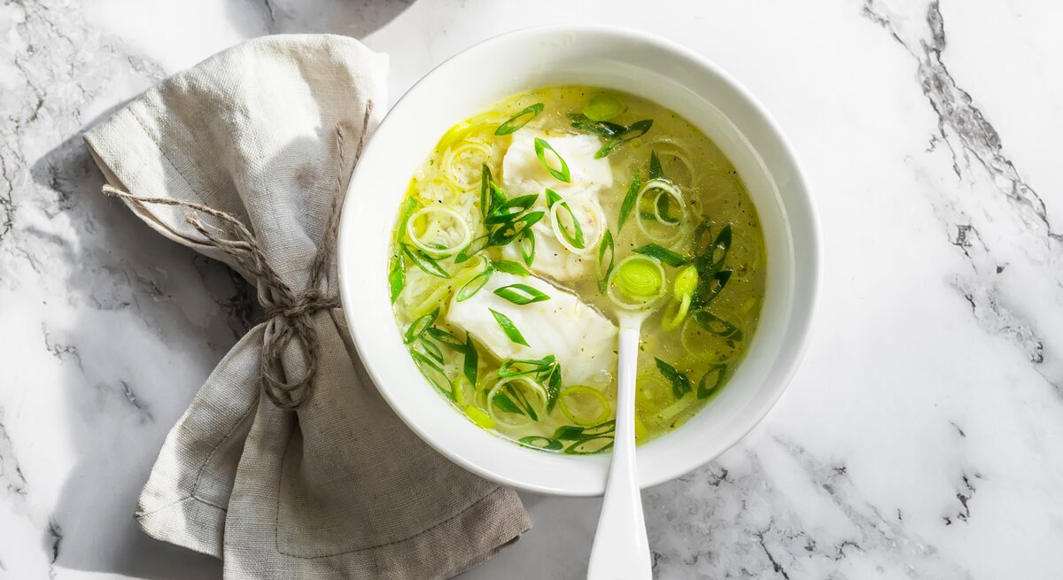 Draufsicht: eine helle Schale mit einer Leichte Lauch-Kabeljau-Suppe steht auf einen Untergrund aus hellem Marmor. In der Suppe ist ein Suppenlöffel.
