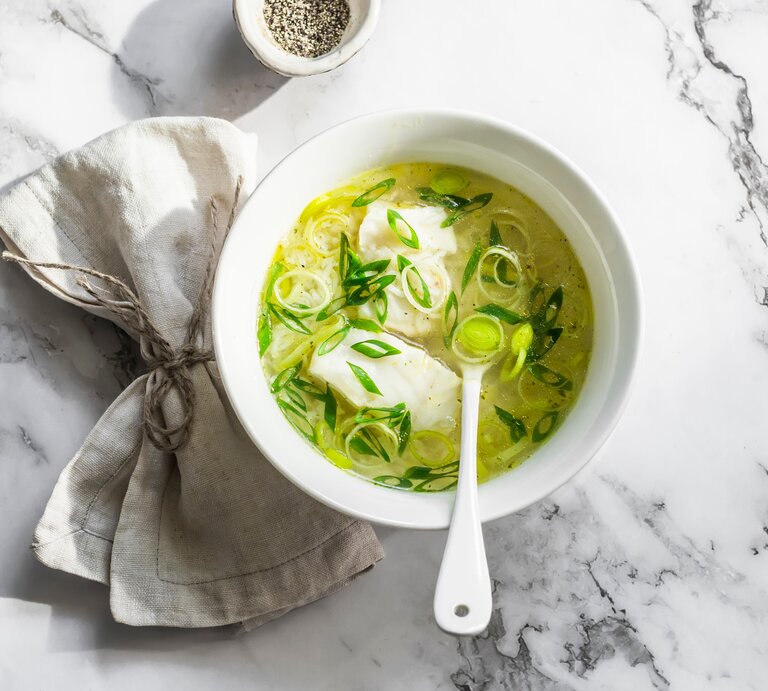 Draufsicht: eine helle Schale mit einer Leichte Lauch-Kabeljau-Suppe steht auf einen Untergrund aus hellem Marmor. In der Suppe ist ein Suppenlöffel. | © Shutterstock / Kiian Oksana