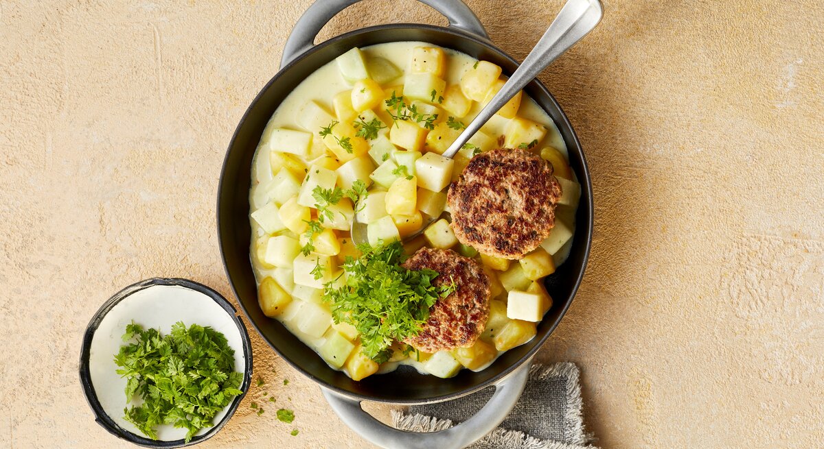 Eine tiefe Pfanne mit den Tatar-Frikadellen mit Kohlrabi-Kartoffel-Ragout, die mit Kerbel garniert sind, neben einer kleinen Schüssel mit Kerbel und einem Tuch, vor einem beigen Hintergrund, von oben. 