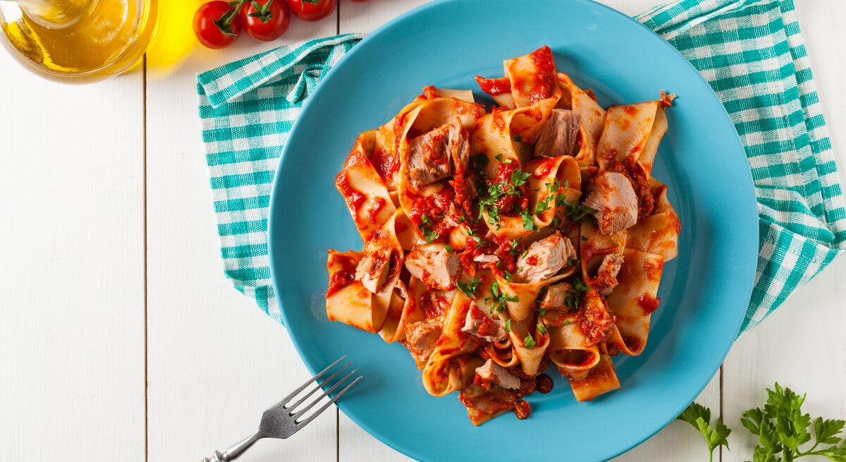 Ein Teller mit einer Portion One-Pot-Pasta mit Thunfisch und Tomatensoße, bestreut mit Petersilie. daneben liegen Tomaten, Petersilienzweige, außerdem ist eine Flasche mit Öl zu sehen.