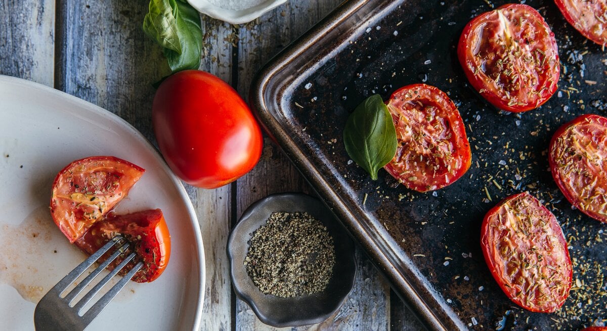 Au der Vogelperspektive: Auf einem Ofenblech liegen halbierte Tomaten, die mit Kräutern betreut sind. Daneben steht ein weißer Teller, auf dem ebenfalls eine halbierte Tomate liegt.